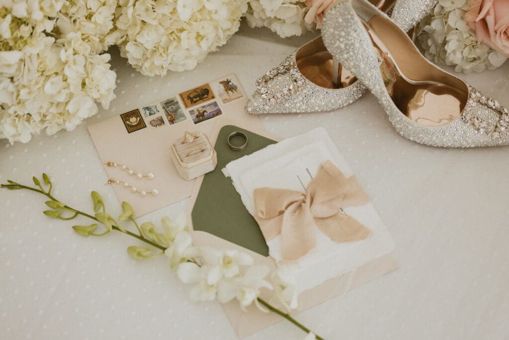 Montage Big Sky wedding invitation suite with custom stationery, silk ribbon, vintage postage, bridal heels, and pearl details styled with white florals.