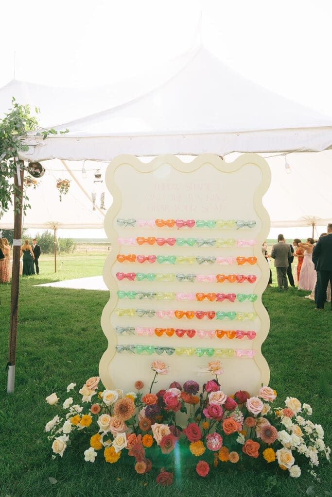 Bright yellow wedding seating chart display with colorful sunglasses escort cards under a white tent at Bozeman, Montana outdoor reception.