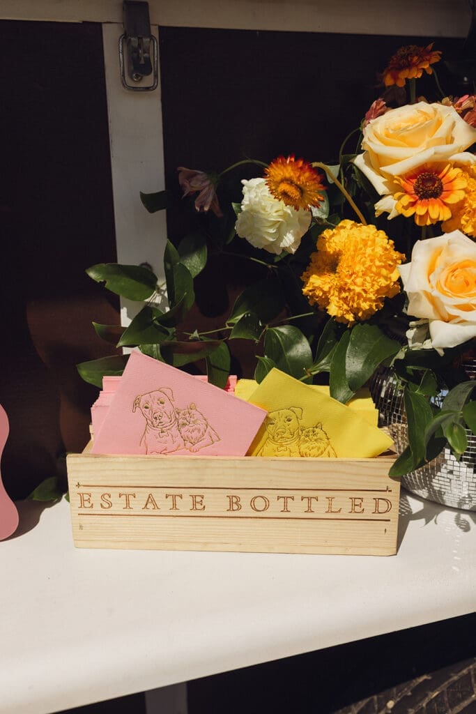 Custom illustrated cocktail napkins featuring the couple’s pets displayed beside vibrant floral arrangement at Montana wedding reception.